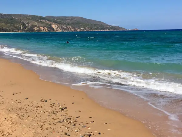 Türkisblaues Meer und ein sandiger Strand bei Oran Stadt. Wellen brechen sanft, im Wasser sind einige Schwimmer zu sehen.